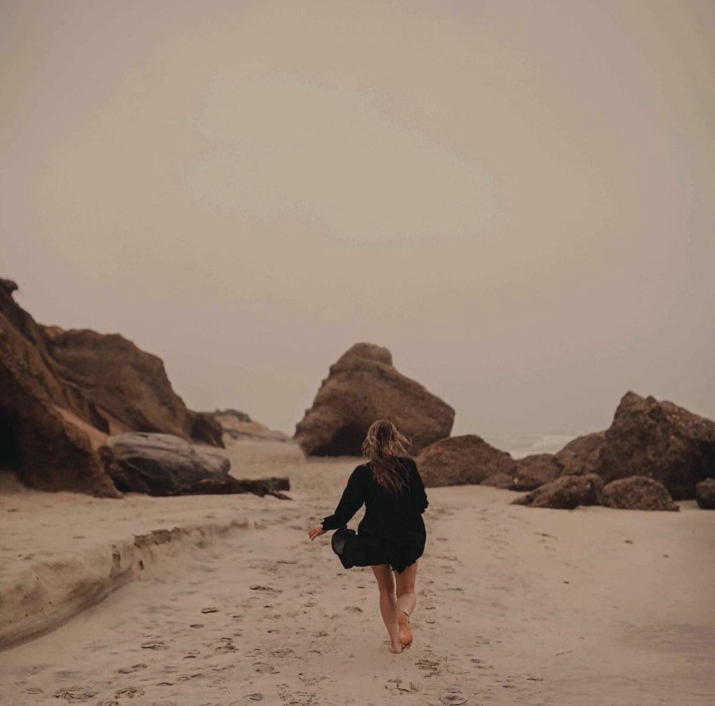 woman black robe running on beach with large rocks